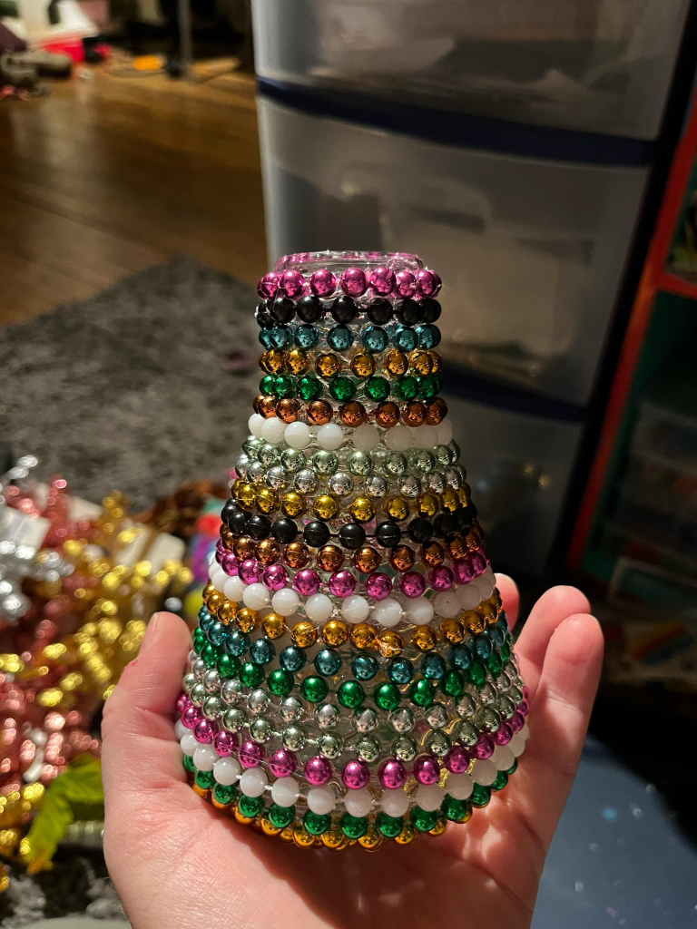 Ashley's hand holds a conical flask with rows of colorful beaded chains glued to the outside. Bead strands are pink, black, blue, gold, green, copper, and white. 